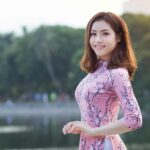 Asian woman in a pink floral dress smiling by a serene lake, surrounded by nature.